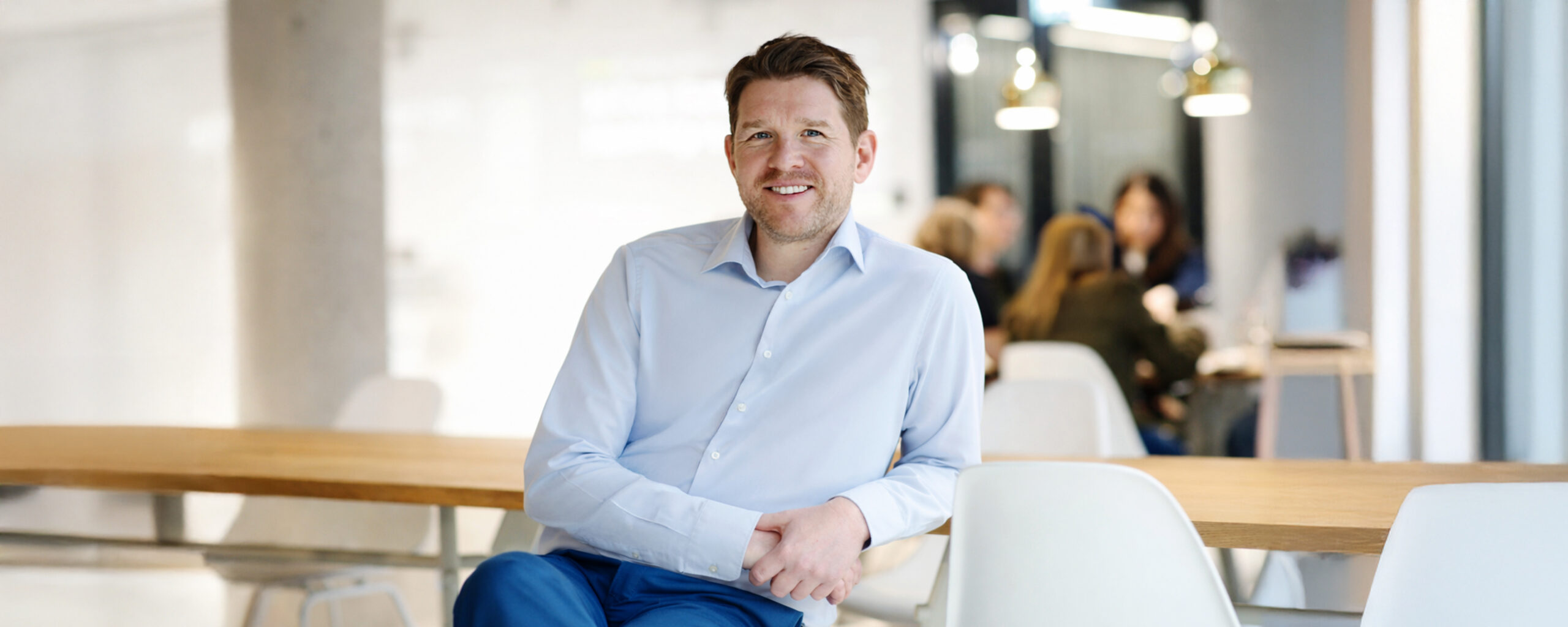 Businessporträt eines Mitarbeiters im hellen Büro mit Kolleginnen und Kollegen im Hintergrund – Mitarbeiterfoto für Unternehmen in Berlin