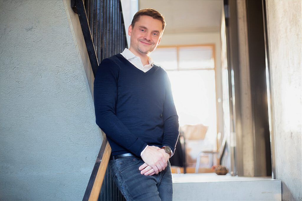 Mitarbeiterfoto eines Mannes im Pullover, lässig an der Wand im Treppenhaus angelehnt – Businessportrait im Unternehmen